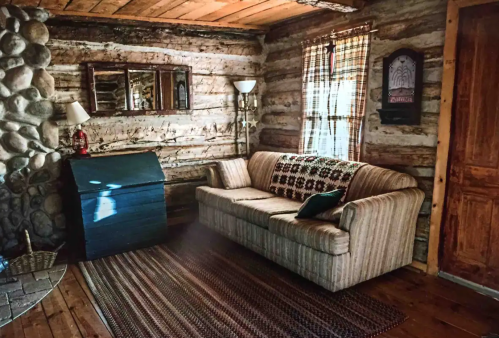 Cozy log cabin interior featuring a striped sofa, wooden walls, a stone fireplace, and a woven rug.